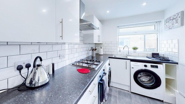 A modern kitchen with white cabinets, a black countertop, a built-in washing machine, and a window with blinds. - Ranworth House - Holiday Home in Bedford, Sleeps 5 (Bedford)