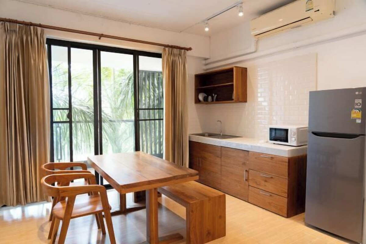 A kitchen with a wooden table, chairs, a microwave, a refrigerator, and a window with curtains.
