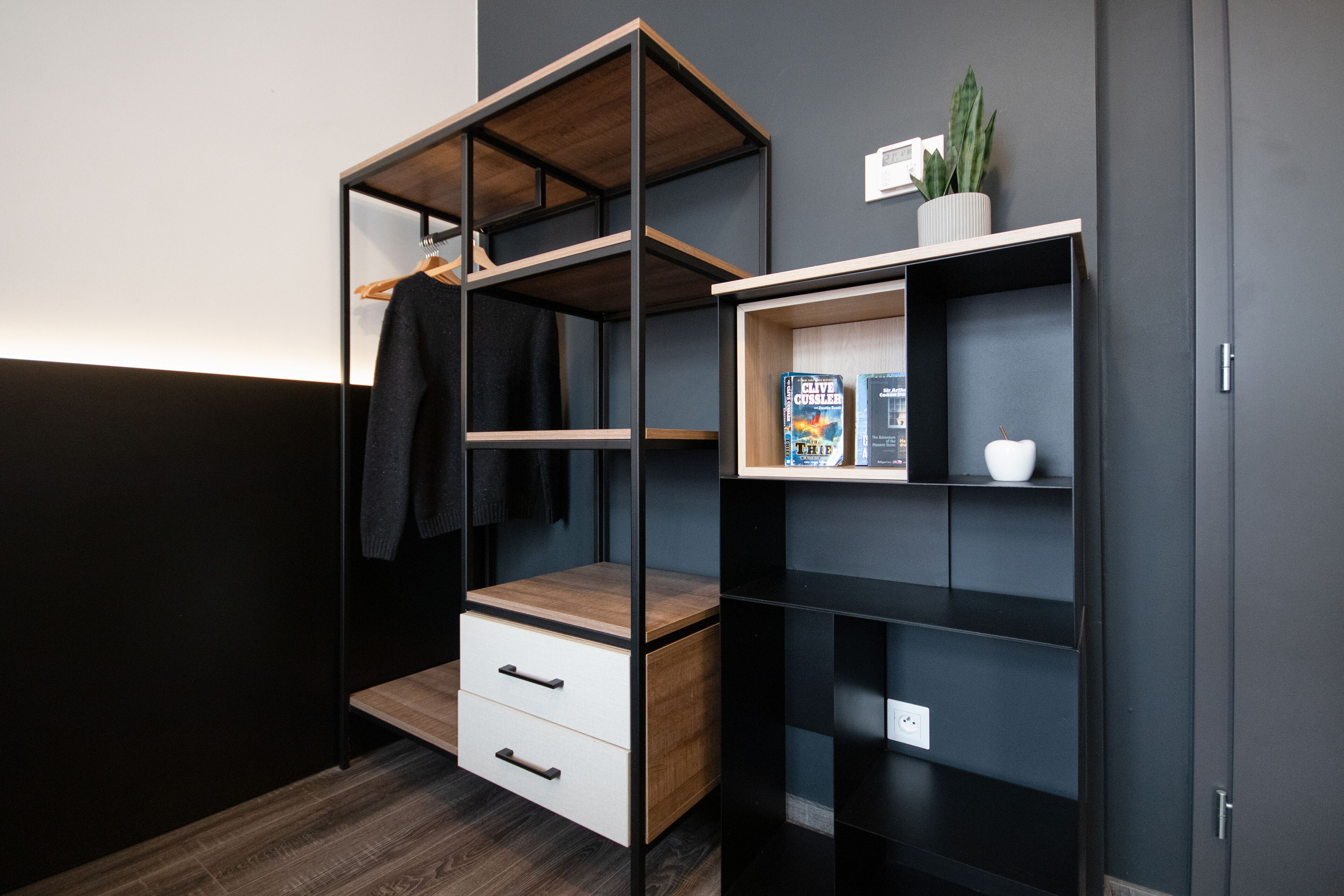 A modern, multi-tiered shelving unit with a wooden top, a white drawer, and a small shelf with a plant and a book.