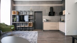 A modern living room with a green armchair, a black TV on a wooden shelf, and a kitchen area with a microwave and cabinets. - CORU House (Prague)