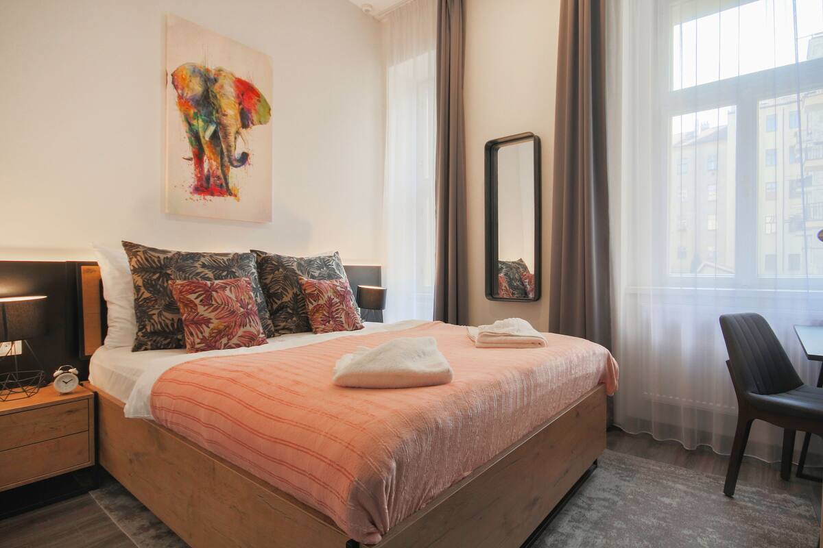A hotel room with a large bed, a nightstand with a lamp, a painting of an elephant, a mirror, and a view of buildings through the window.