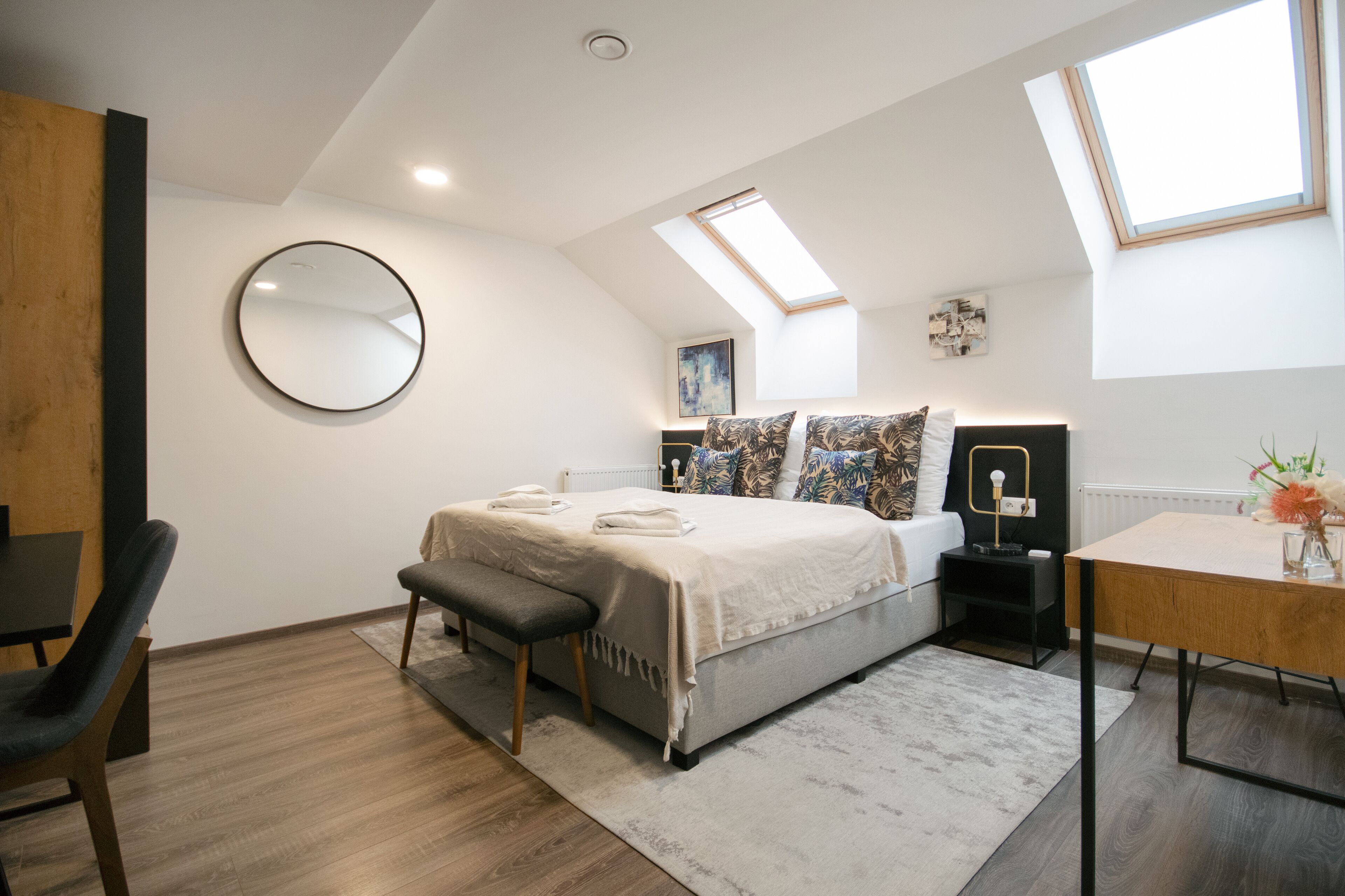 Un dormitorio moderno con tragaluz, un espejo redondo, cama con banqueta, una mesita y una silla.