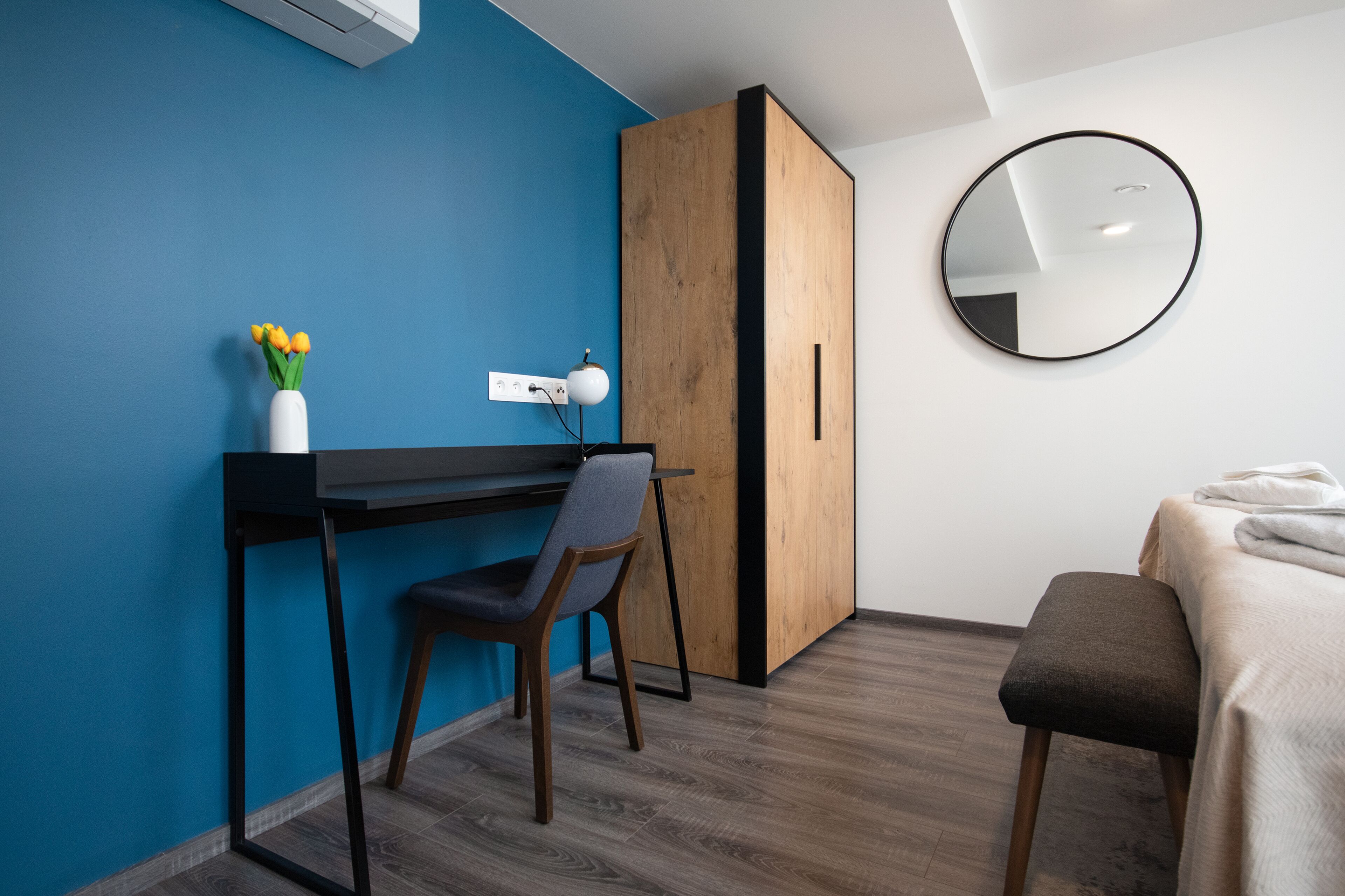Une chambre moderne avec un mur d’accent bleu, une armoire en bois, un miroir rond, un bureau avec une chaise et un lit recouvert d’une couverture blanche.