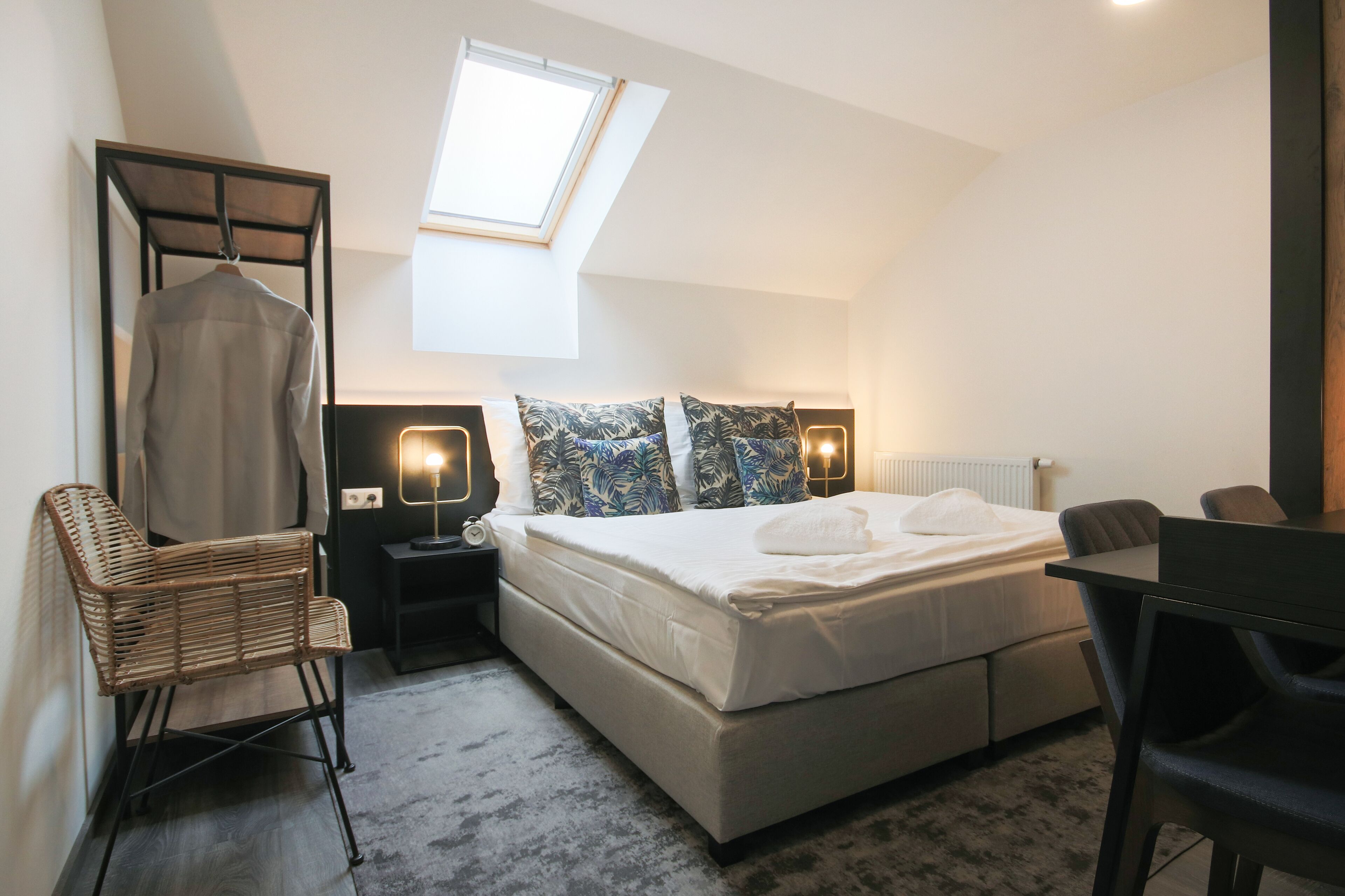 A bedroom with a bed, two nightstands, a desk, a chair, a hanging rack, a window, and a radiator.