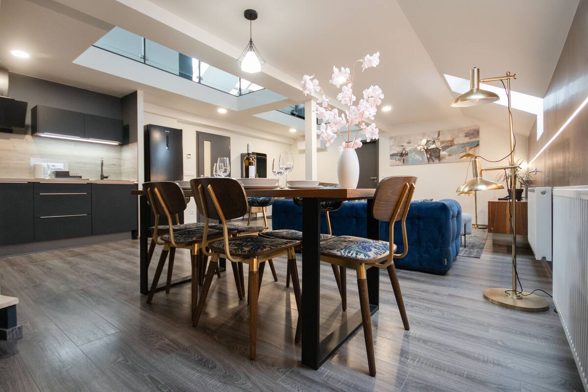 A modern kitchen with dark cabinetry, a dining area with a wooden table and chairs, and a blue sofa in a living area with a large window and skylight.