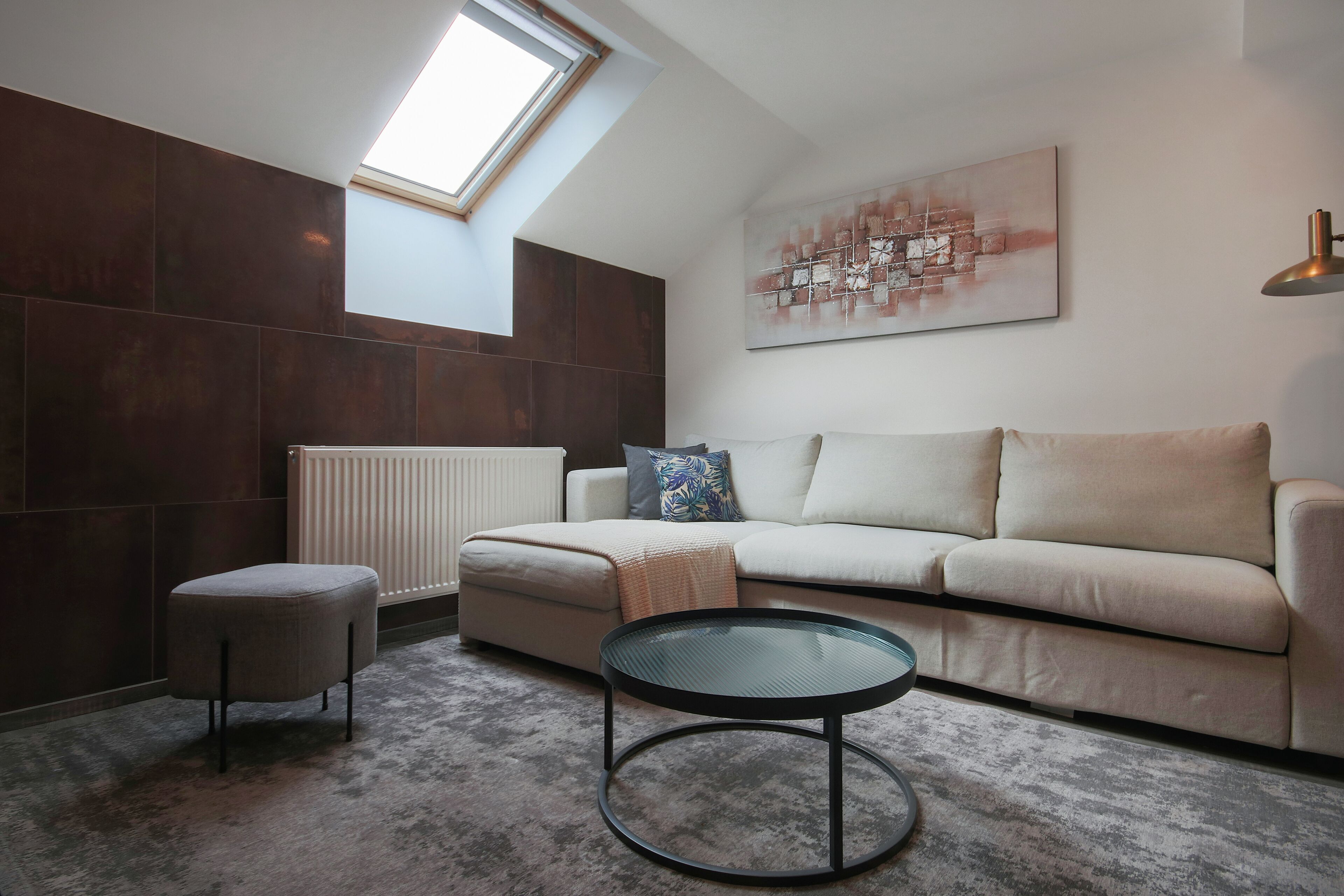 Un salon moderne avec un plafond haut, une lucarne, un canapé beige, une table basse en verre et un radiateur.