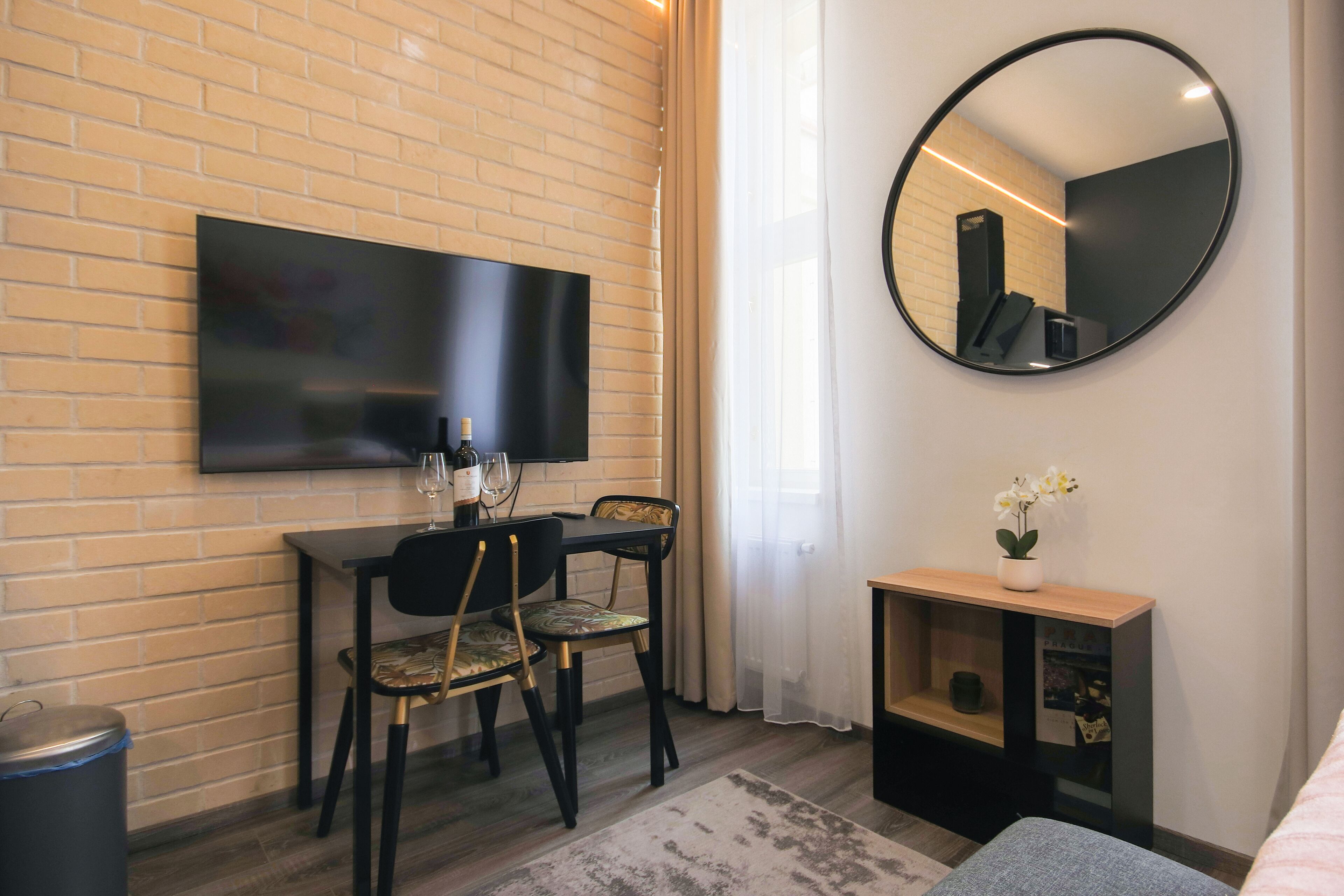 Quarto moderno com parede de tijolos, televisão de tela plana acima de uma mesa de jantar com cadeiras, espelho redondo e pequena mesa de madeira com uma planta.
