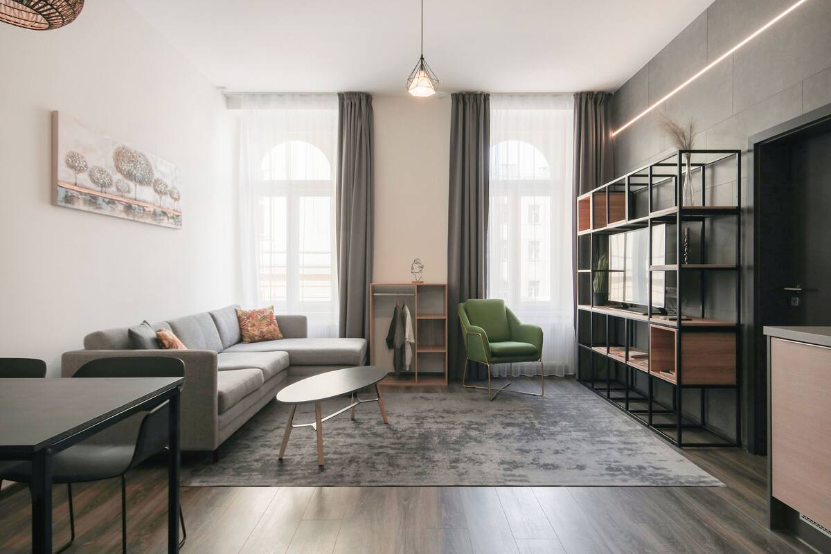 A modern living room with a sofa, coffee table, and a green armchair. There is a bookshelf and a dining area with a table and chairs.