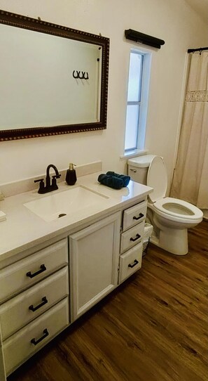 Un baño con lavamanos blanco, grifo negro y un inodoro. Hay una ventana sobre el inodoro y un espejo sobre el lavamanos.