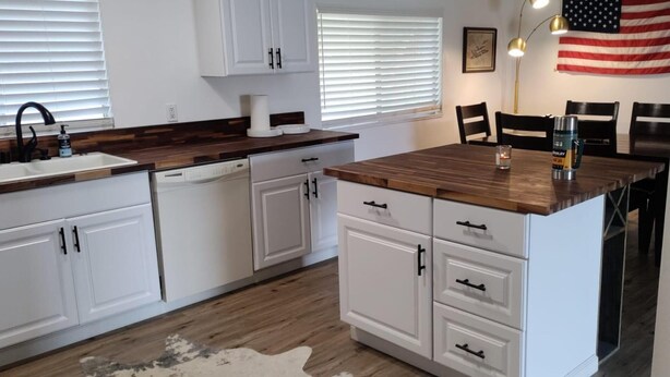 A modern kitchen with white cabinets, a wooden countertop, and a dishwasher. There is a dining area with a table and chairs, and an American flag on the wall. - Cozy Lake House Surrounded by Mountain's (Wofford Heights)
