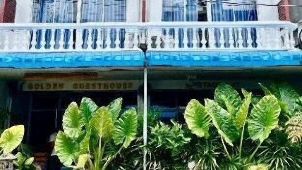 Un immeuble à plusieurs étages doté de balcons, une devanture visible avec une enseigne, et une végétation luxuriante à l’avant.