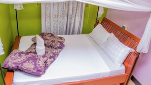 Una cama individual con colchón blanco, manta morada y toalla blanca. La cama tiene cabecera de madera y está apoyada contra una pared verde.