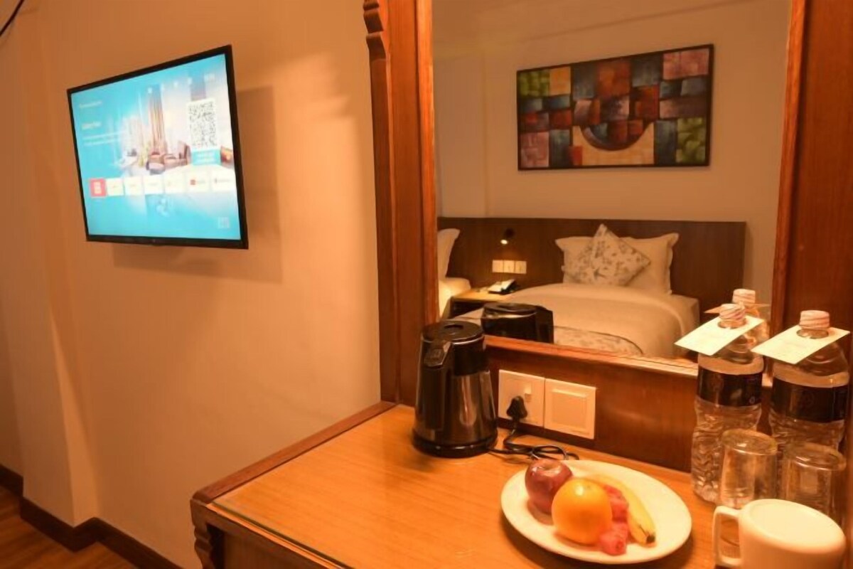 A hotel room with a bed, a bedside table with a coffee maker, a plate of fruit, and a TV mounted on the wall.