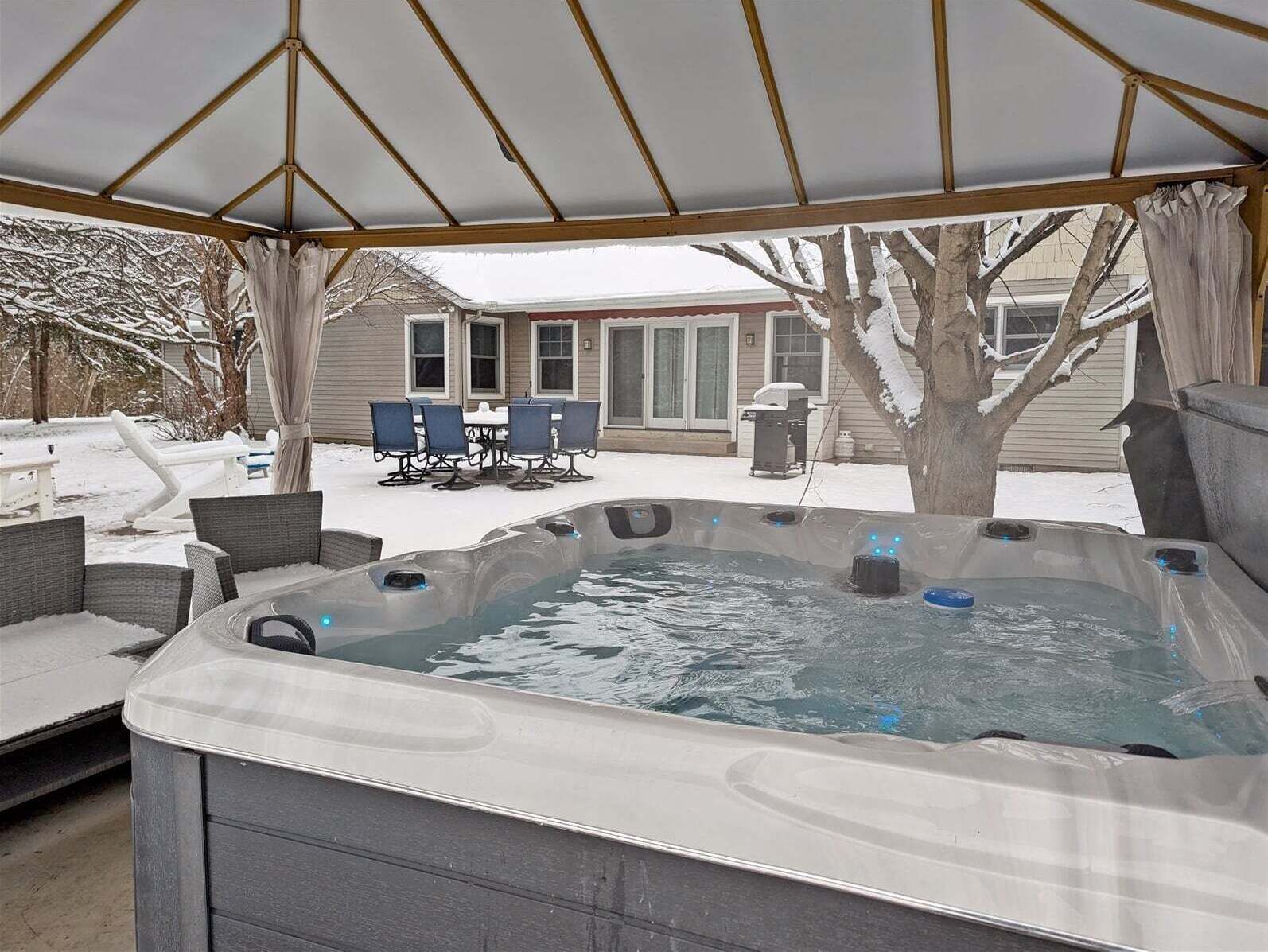 Een hottub onder een overdekt terras met een tafel en stoelen, omringd door sneeuw.