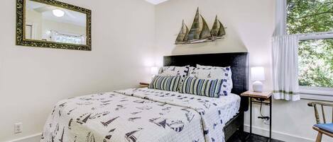 A bedroom with a bed, a nightstand, a lamp, a chair, a window, and a decorative ship model on the wall.