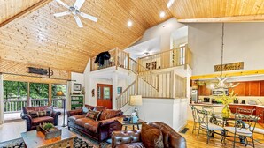 A spacious living room with leather sofas, a dining area with metal chairs, a wooden staircase, and a kitchen with wooden cabinets.