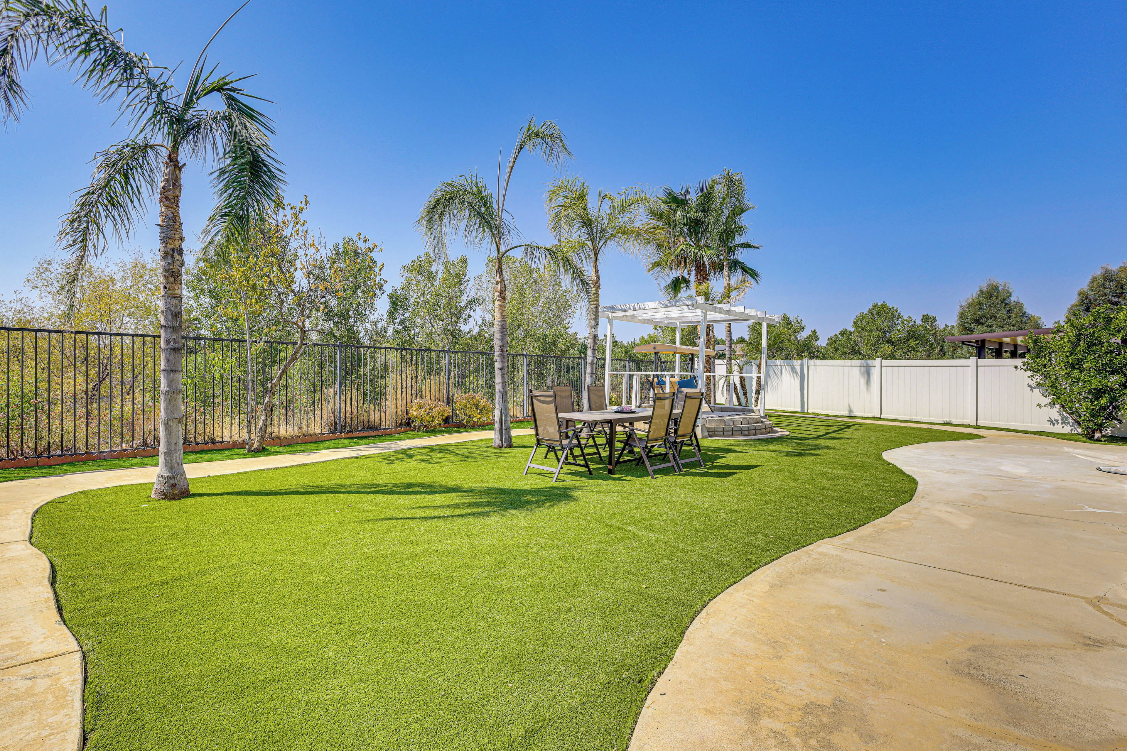 Un patio trasero con césped artificial, una mesa con sillas, una pérgola y palmeras.