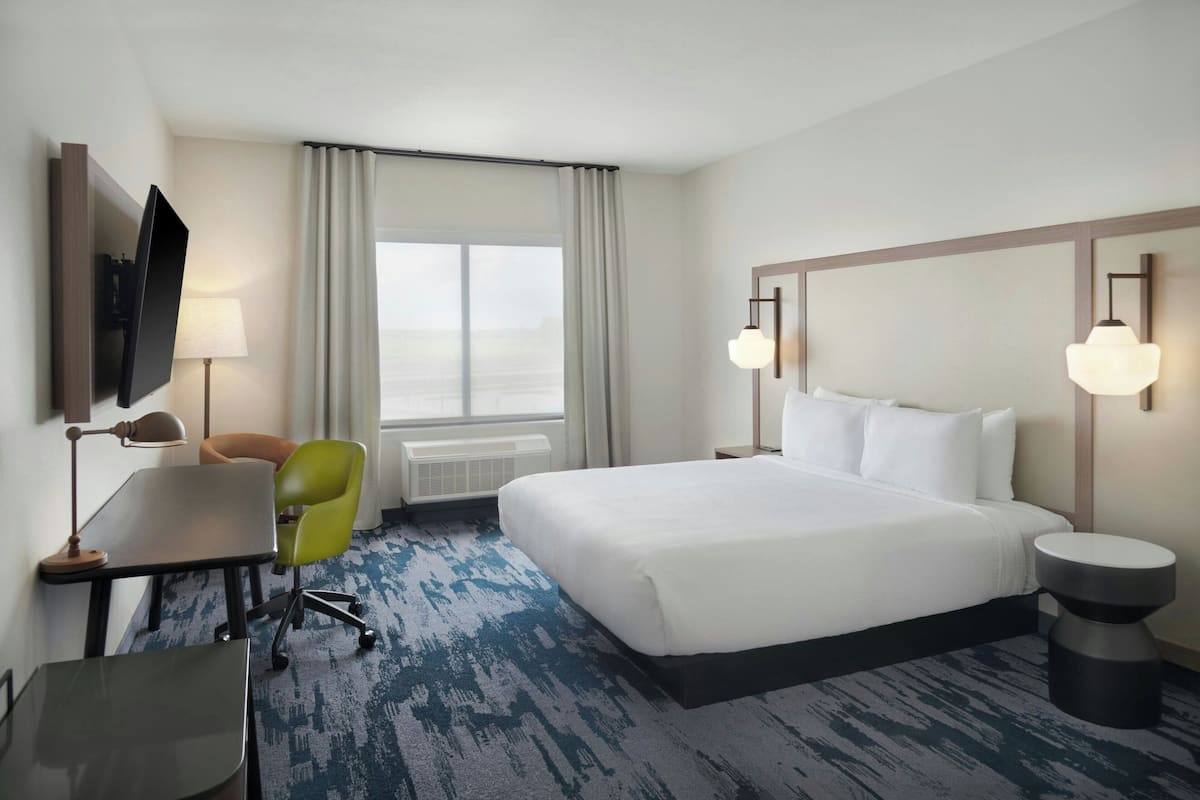 A hotel room with a large bed, a desk with a chair, a TV mounted on the wall, and a window with curtains.