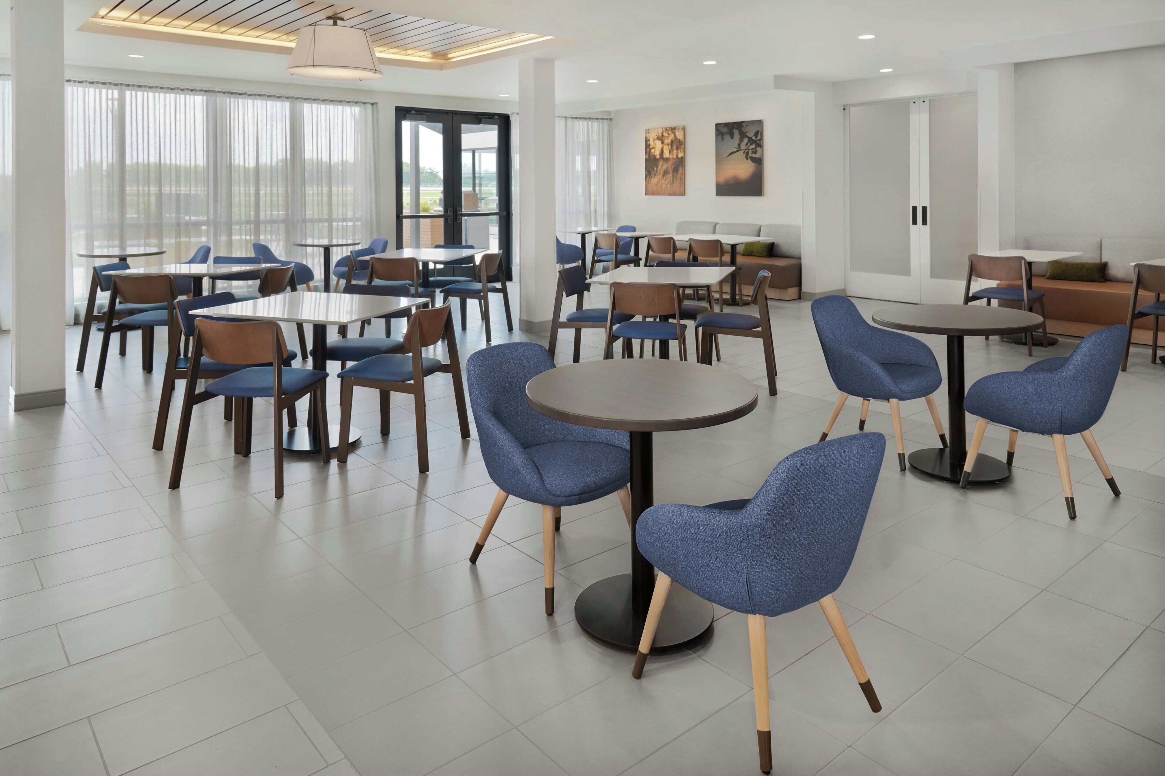Comedor moderno de hotel con mesas redondas y sillas azules, ventanas amplias y obras de arte en las paredes.