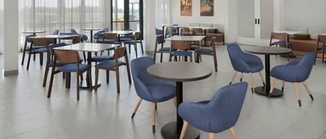 Ein moderner Hotel-Speisesaal mit runden Tischen und blauen Stühlen, großen Fenstern und Kunstwerken an den Wänden.