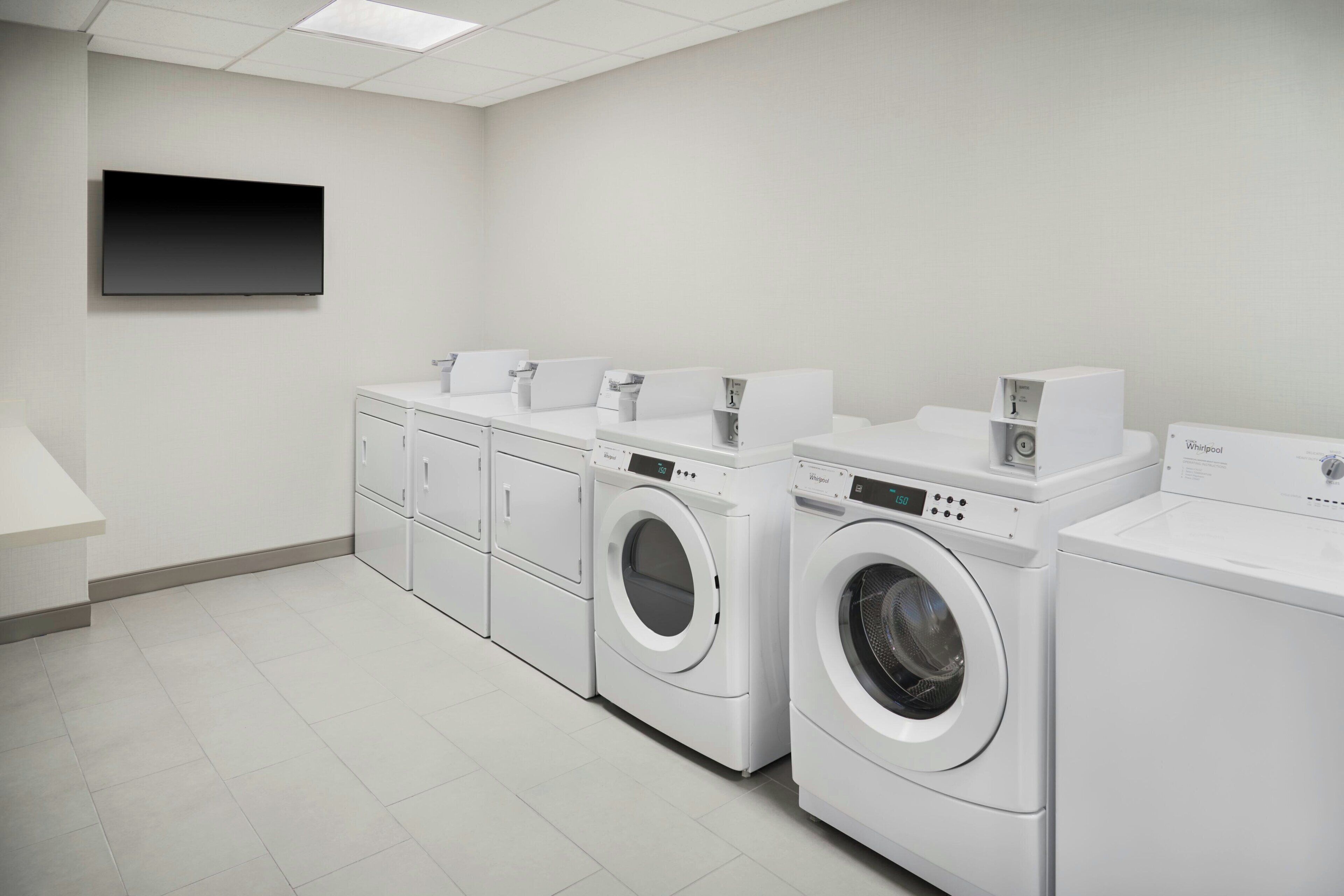 A laundry room with three front-loading washing machines and a dryer, a flat-screen TV mounted on the wall, and a white tiled floor.