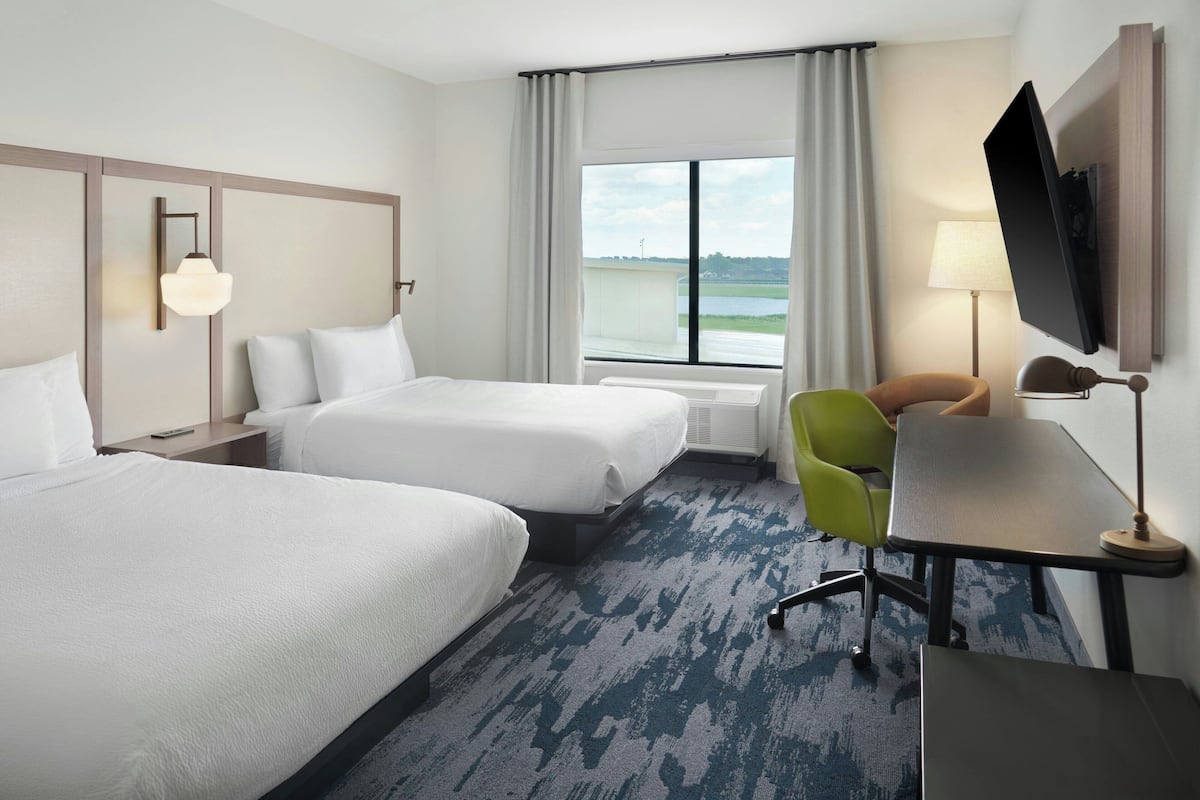 A hotel room with two beds, a desk, a chair, a TV, and a window with a view of the outdoors.