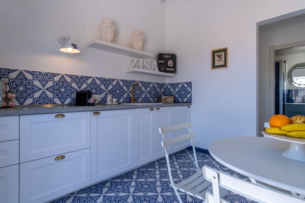 Una cocina con armarios blancos, salpicadero con estampado azul y una mesa redonda con fruta.