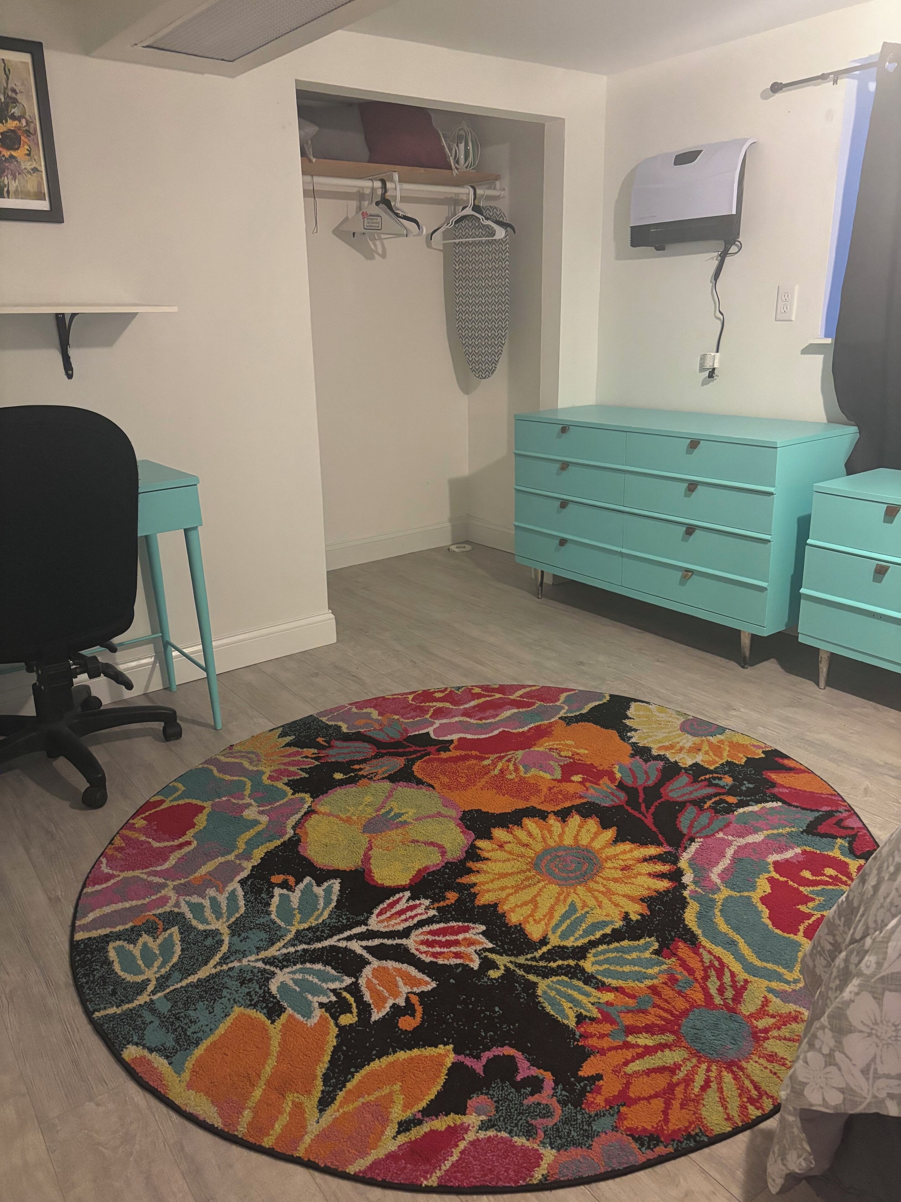 Una habitación con un escritorio color turquesa, una silla, un aparador color turquesa, un tapete con motivos florales coloridos y un aire acondicionado de pared.