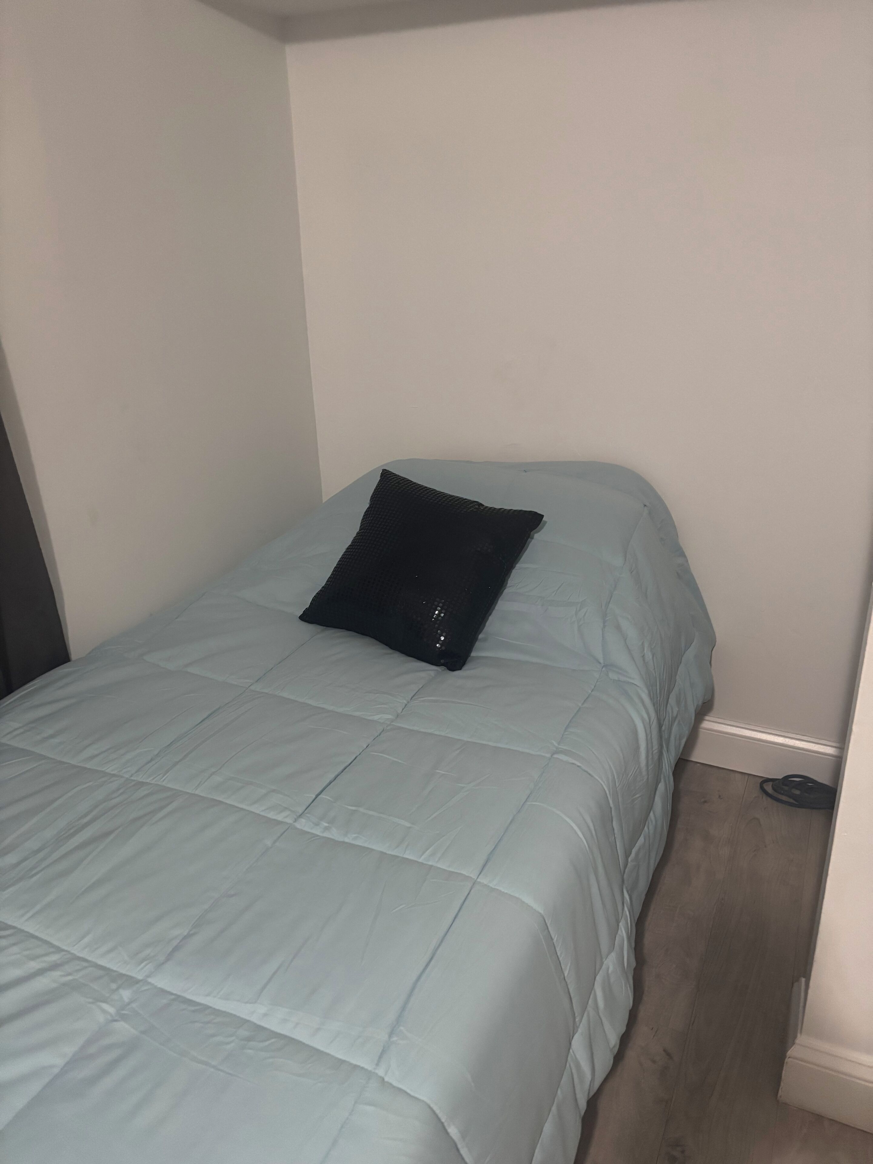 Een enkel bed met een lichtblauwe dekbedovertrek en een zwart kussen in een kamer met witte muren en een houten vloer.