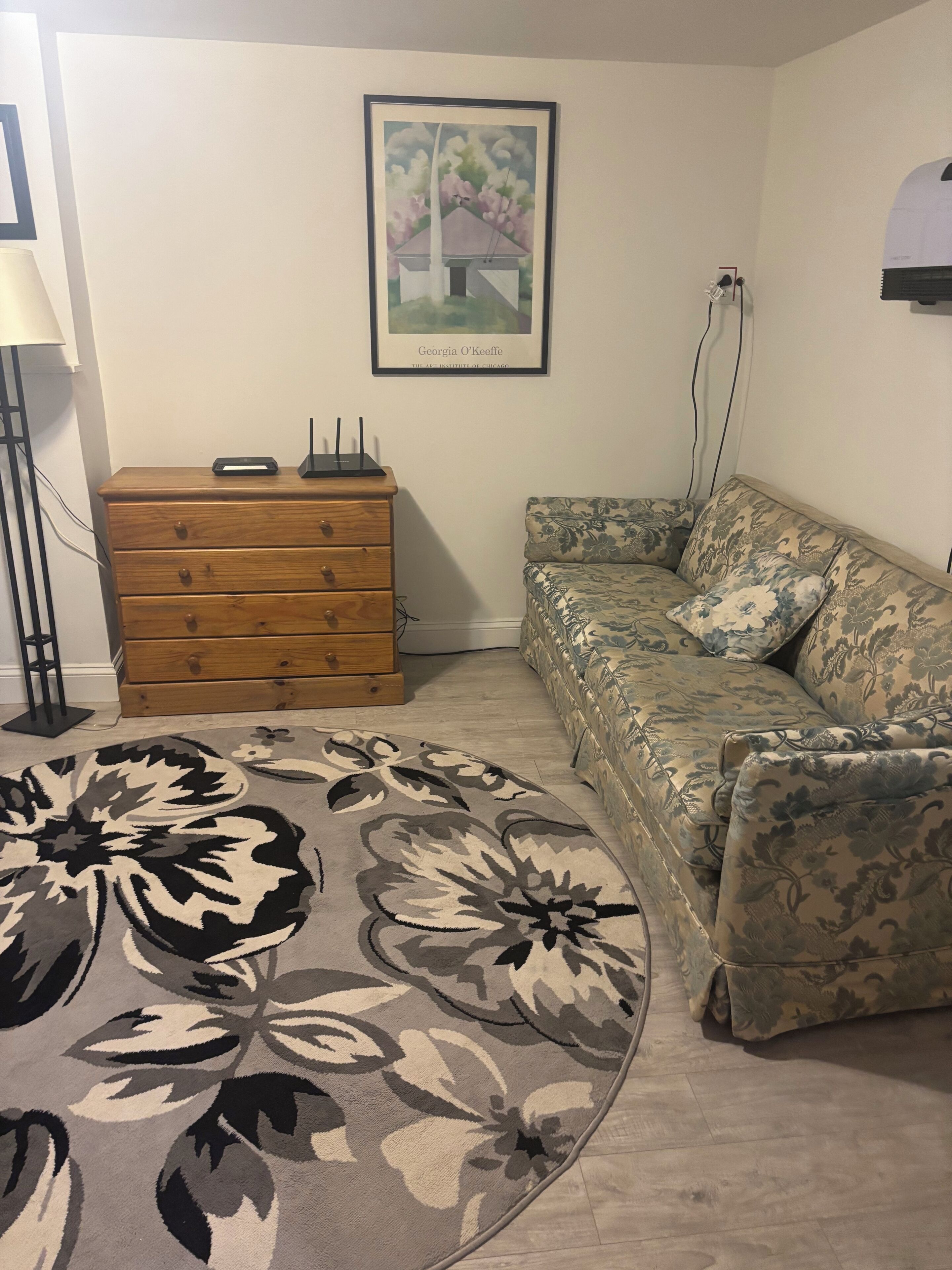 A room with a wooden dresser, a floral-patterned sofa, a framed artwork on the wall, and a lamp.