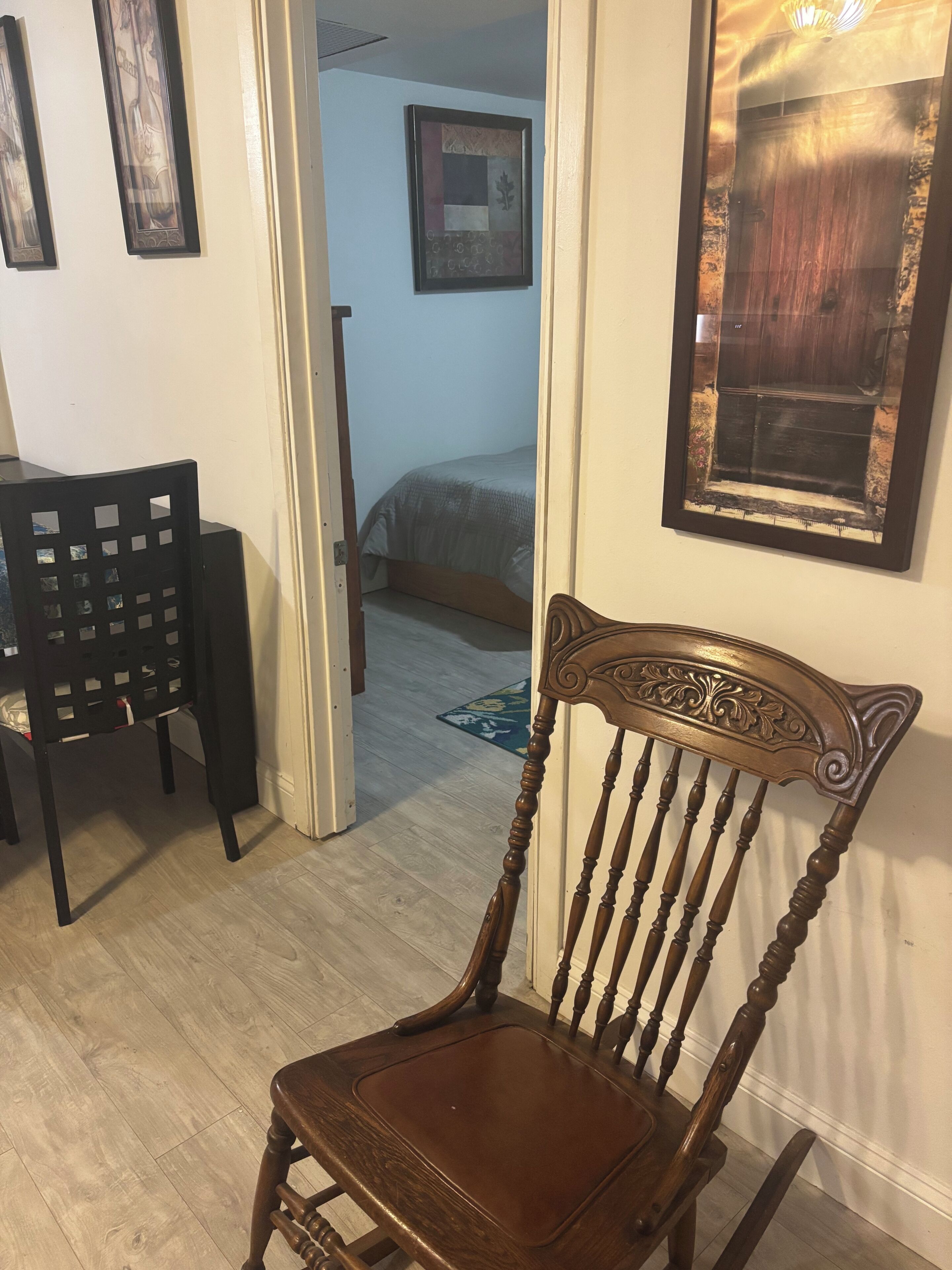 Una silla de madera con asiento y respaldo acolchados, una mesa de madera con una silla negra, y una cama con colcha gris al fondo.