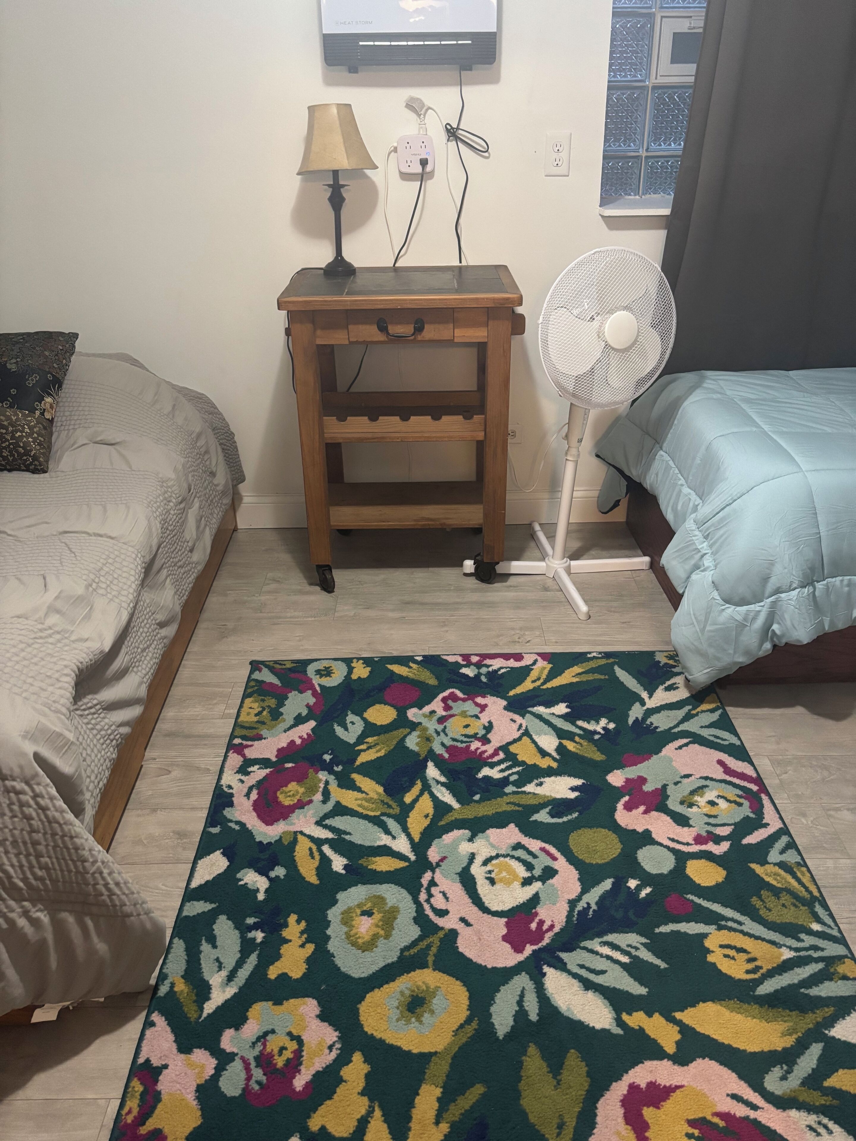 Une chambre à coucher avec un lit, une table de chevet, une lampe, un ventilateur et une fenêtre avec un rideau.