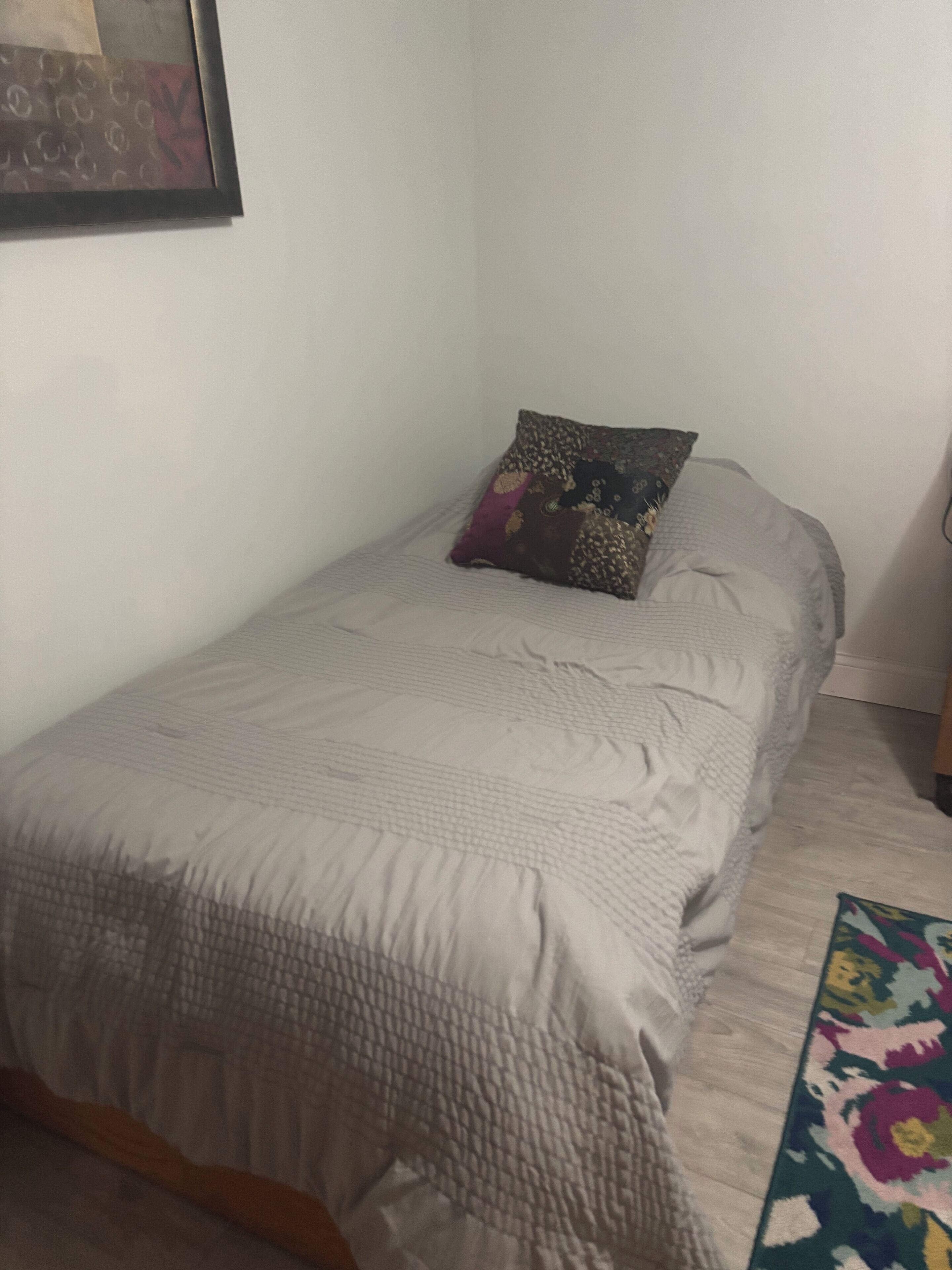 A single bed with a patterned pillow, a plain wall, and a wooden floor.