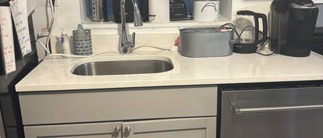 Een keuken met een spoelbak, aanrecht en kastjes. Er is een raam met blauw getinte glas, een koffiezetapparaat en een waterkoker.