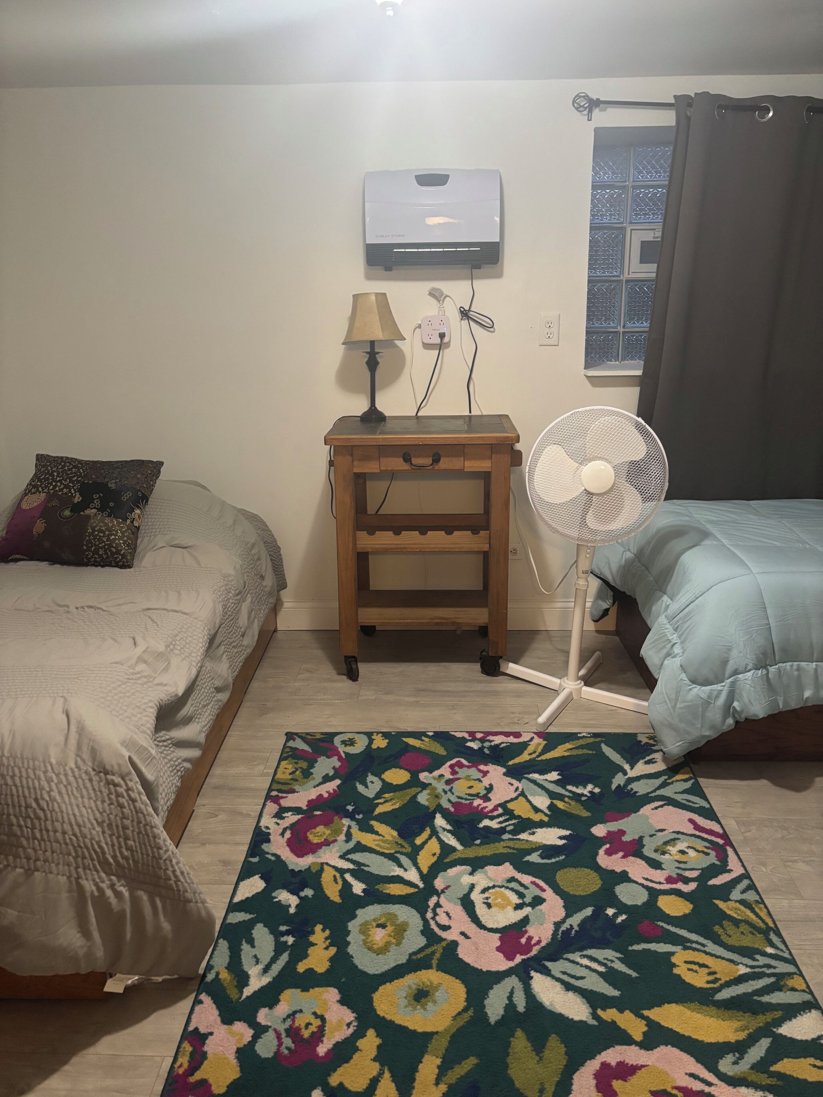 Un dormitorio con dos camas, una mesita de noche con lámpara, un ventilador de pie y un aire acondicionado en la pared.