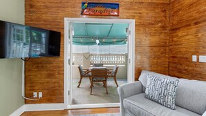 A room with wooden walls, a television mounted on the wall, a sofa with a patterned cushion, and a dining area with a table and chairs. - Poolside Hide-Away @ Palm Gardens offered by Rent Key West Vacations (Key West)