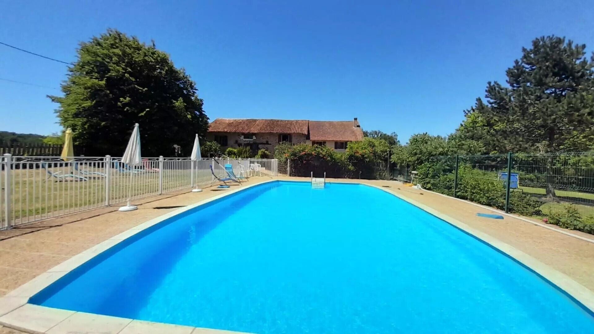 Une piscine rectangulaire à l’eau d’un bleu limpide, entourée d’une clôture blanche. Des chaises longues et des parasols sont disposés le long du bord de la piscine.