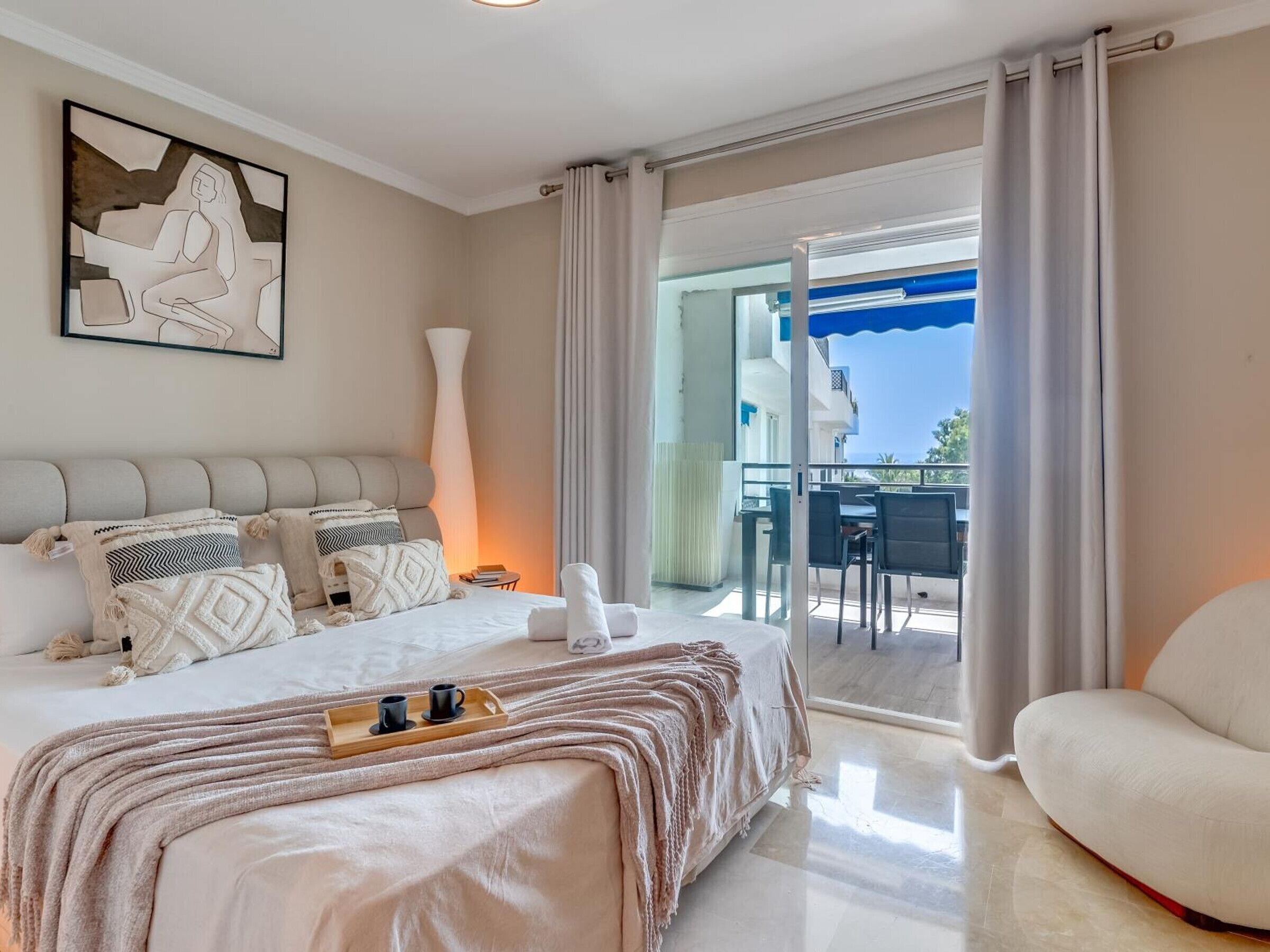 A modern hotel room with a large bed, a balcony with a table and chairs, and a view of the sea.