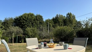 Un espace extérieur aménagé pour les repas, avec une table ronde, des chaises et, au fond, une balançoire.