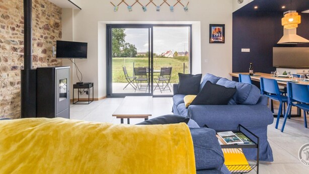 A modern living room with a sofa, a yellow ottoman, and a dining area with blue chairs.