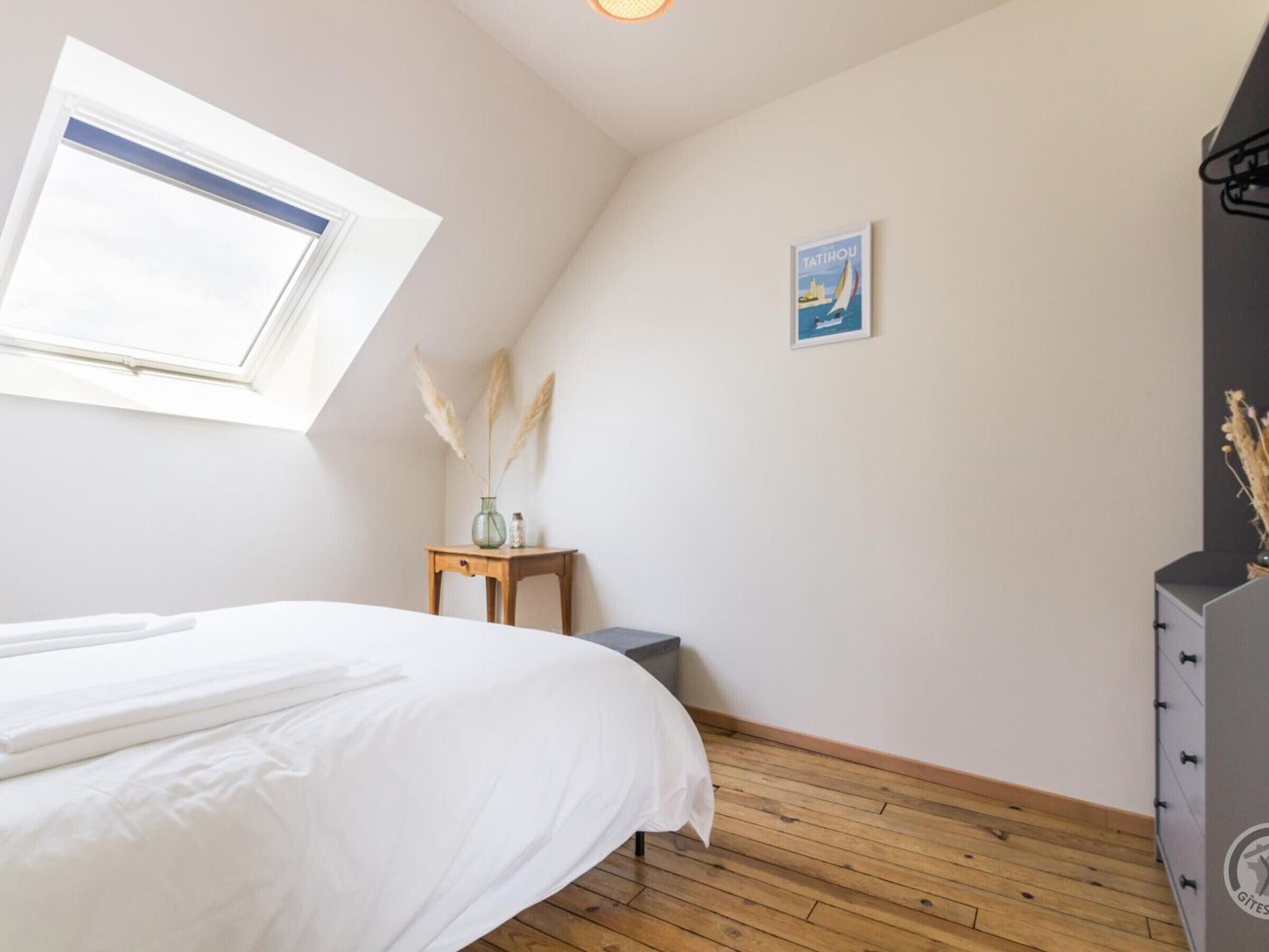 Een slaapkamer met een bed, een klein tafeltje met een vaas, een lijst met een foto aan de muur, en een dressoir.