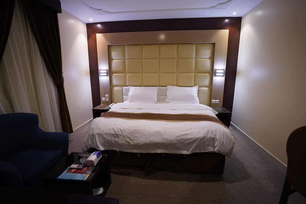 Une chambre d’hôtel avec un grand lit, deux tables de chevet, un fauteuil bleu et une petite table avec un magazine.