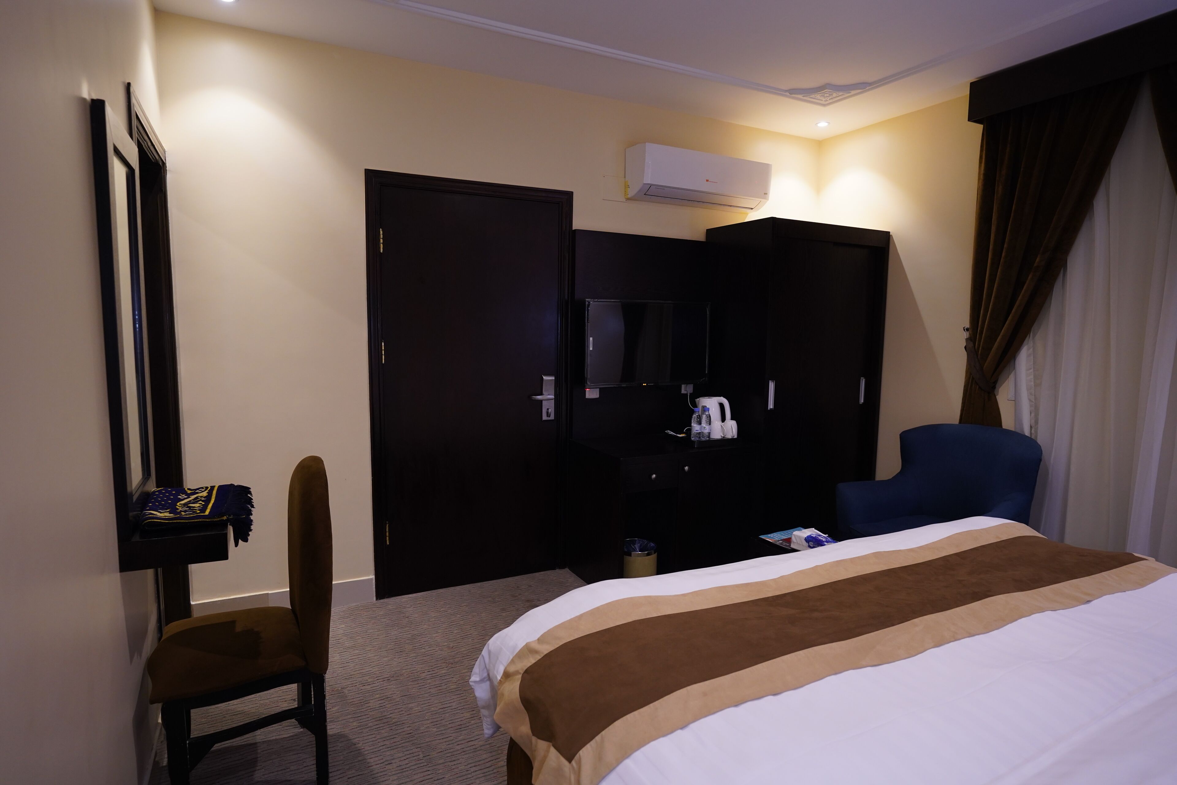 A hotel room with a bed, a desk, a chair, a wardrobe, and an air conditioning unit.