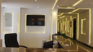 Zona de recepción de un hotel con mostrador de mármol, una silla de oficina negra, un televisor de pantalla plana montado en la pared y un pasillo con paredes y puertas iluminadas.