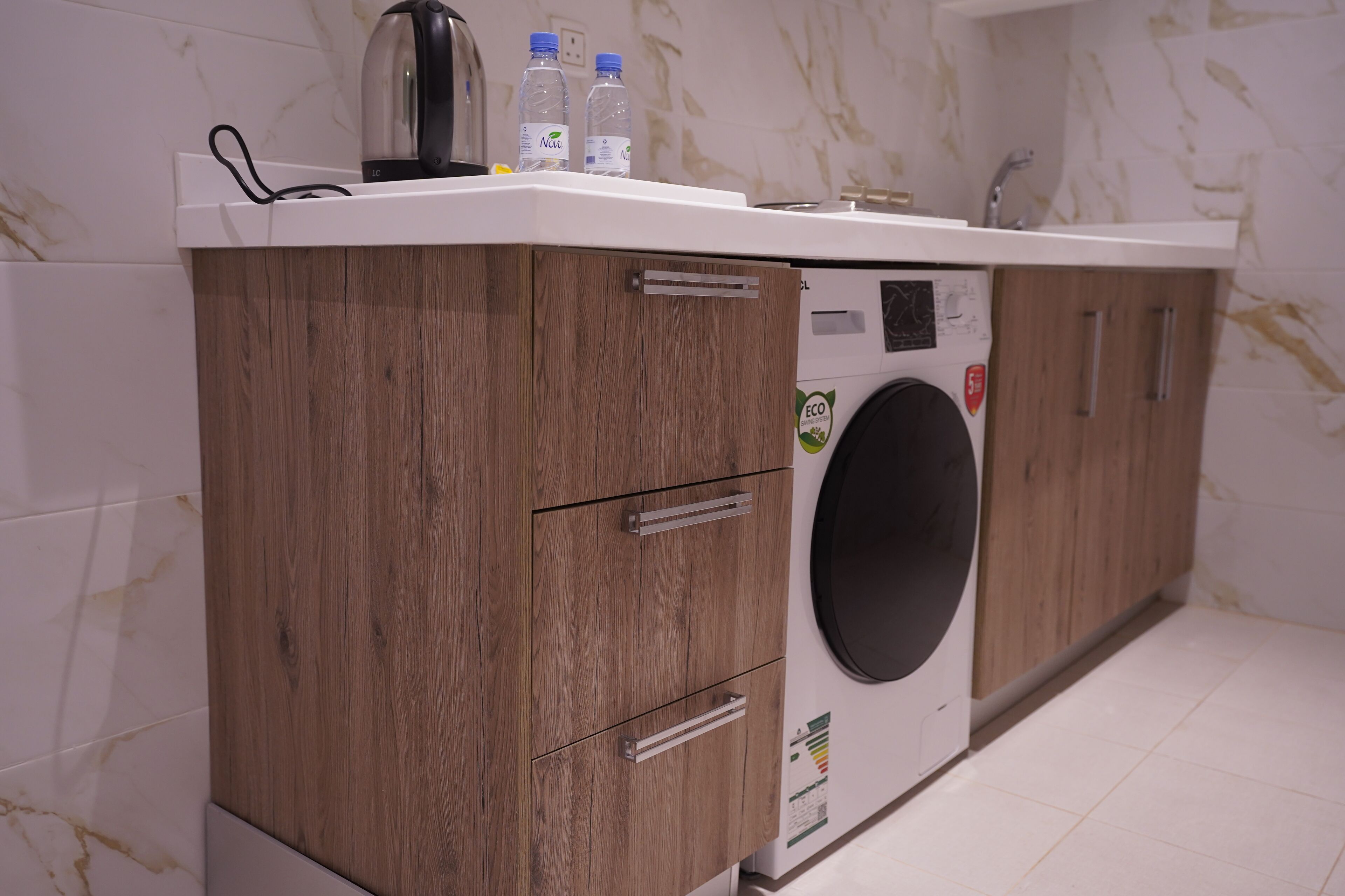 A compact laundry area with a washing machine, wooden cabinets, and a countertop with a kettle and water bottles.