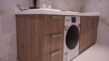 A compact laundry area with a washing machine, wooden cabinets, and a countertop with a kettle and water bottles.