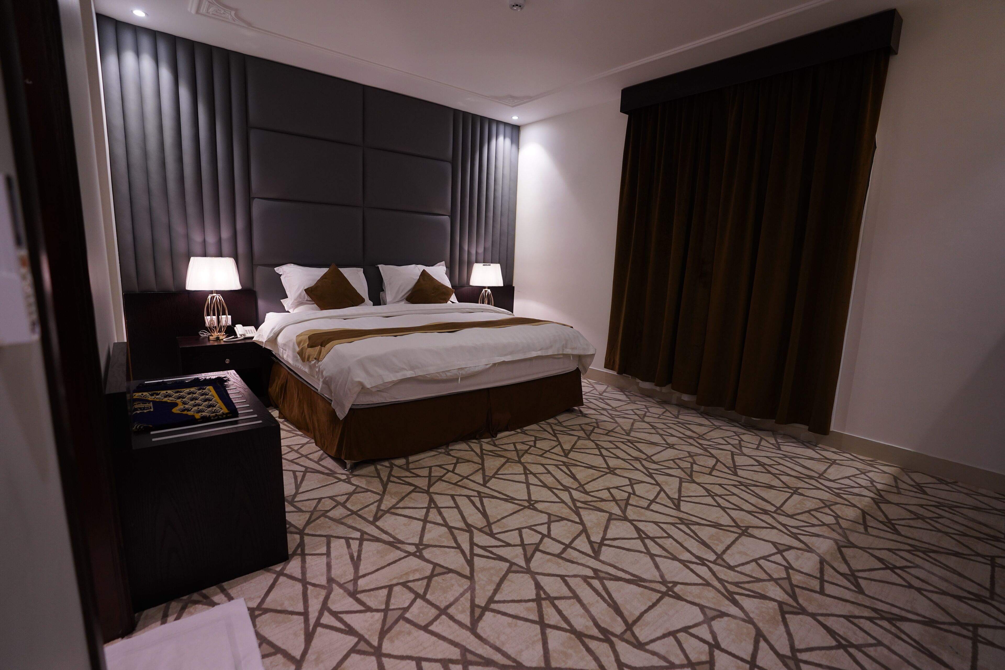 A hotel room with a large bed, dark headboard, patterned carpet, and a nightstand with a lamp.