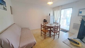 Una stanza piccola e luminosa con un letto singolo, un tavolo da pranzo con sedie, una ventola a soffitto e un frigorifero.