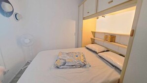 Una camera da letto piccola e ordinata con un letto singolo, un comodino, uno specchio fissato alla parete e una ventola a soffitto.
