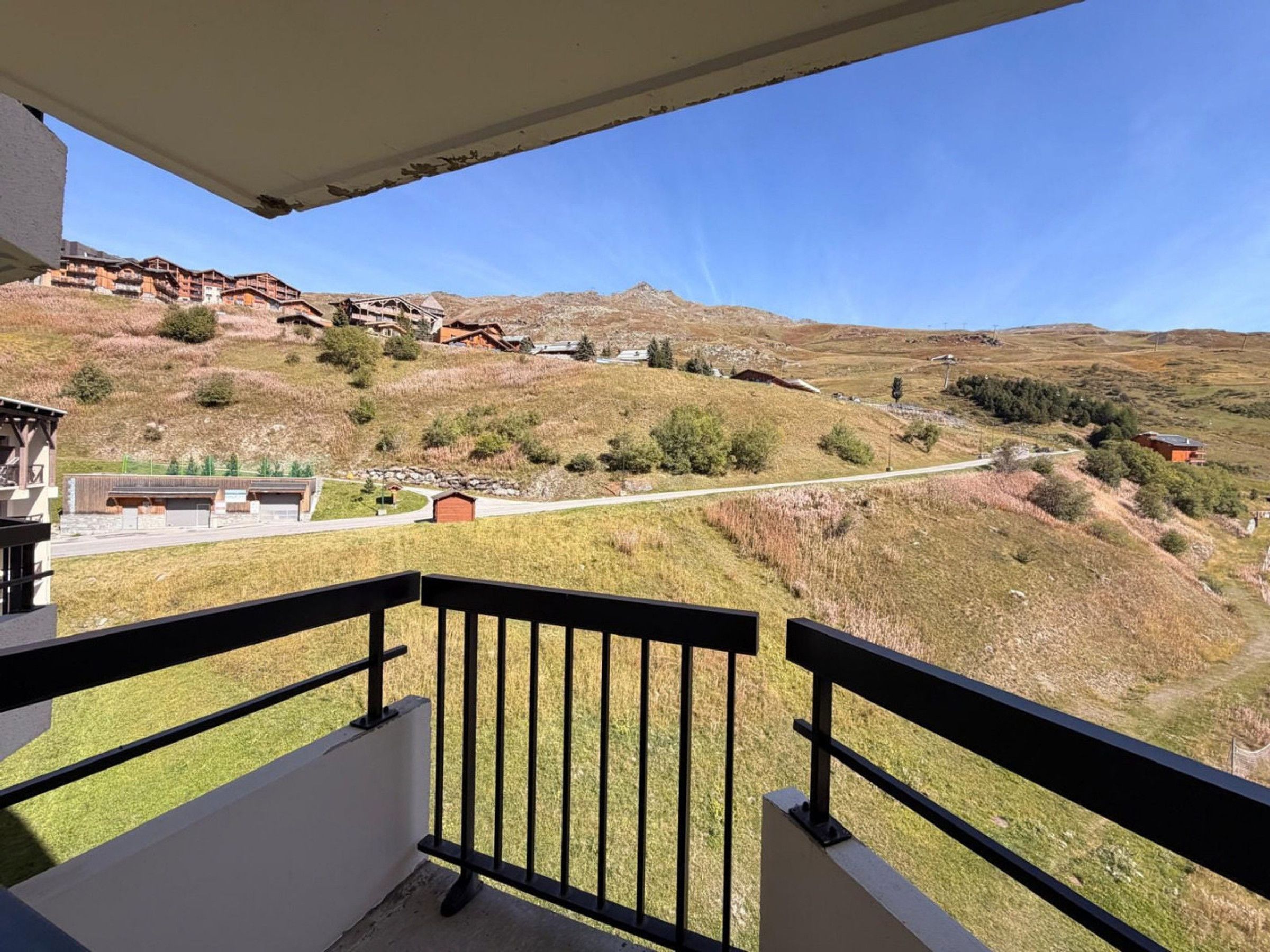 A balcony with a view of a hillside, a building with a sloped roof, and a clear sky.